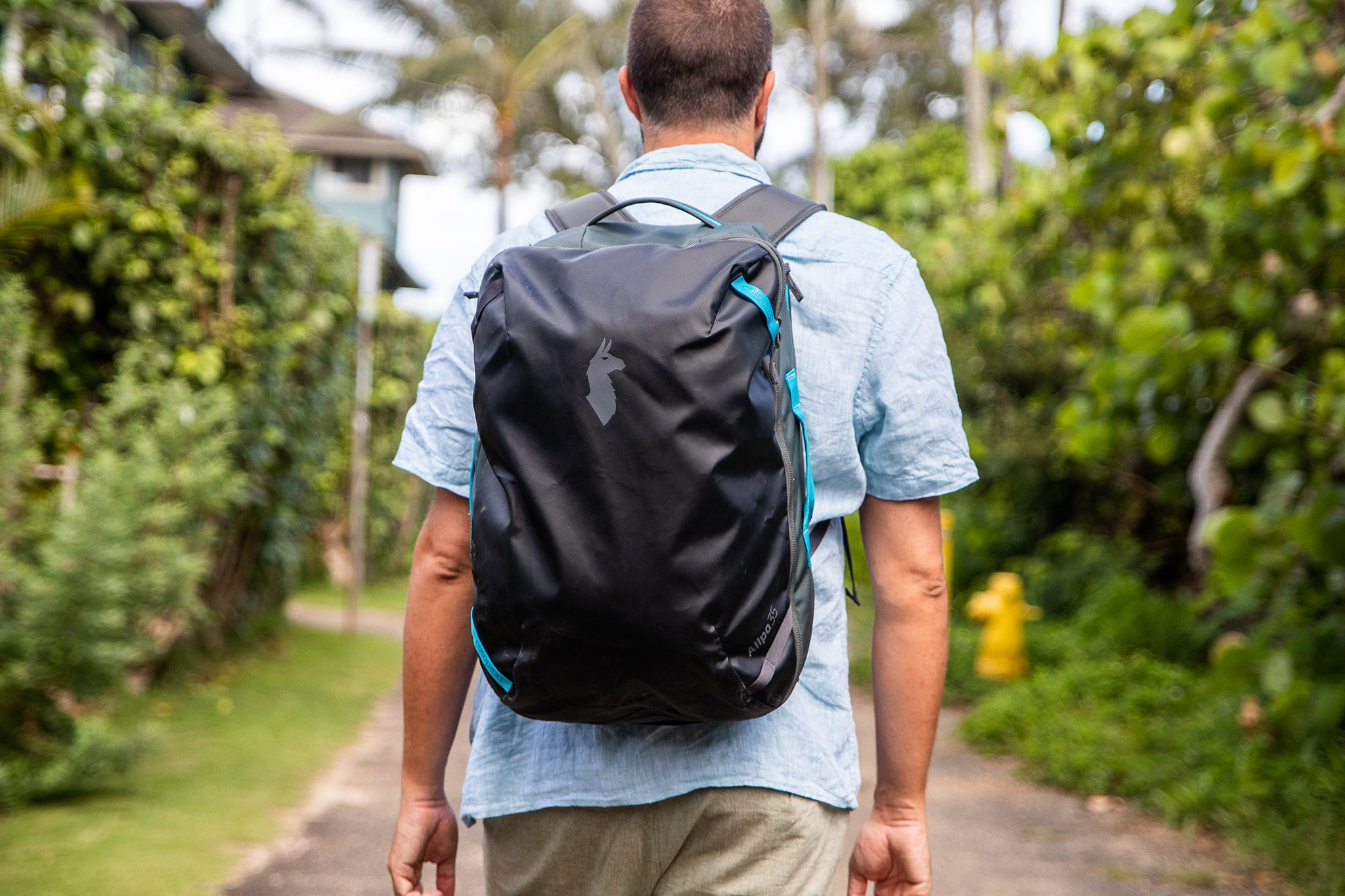 Ba lô Cotopaxi Allpa 35L màu đen với logo đầu lạc đà không bướu màu xám và các điểm nhấn màu xanh nước biển, người đang mặc áo phông ngắn tay màu xanh nhạt.