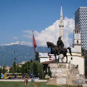 Tirana, with a statue of a man on a horse, a mosque with a minaret, a giant blood-red flag, a tall skyscraper, and mountains in the background.