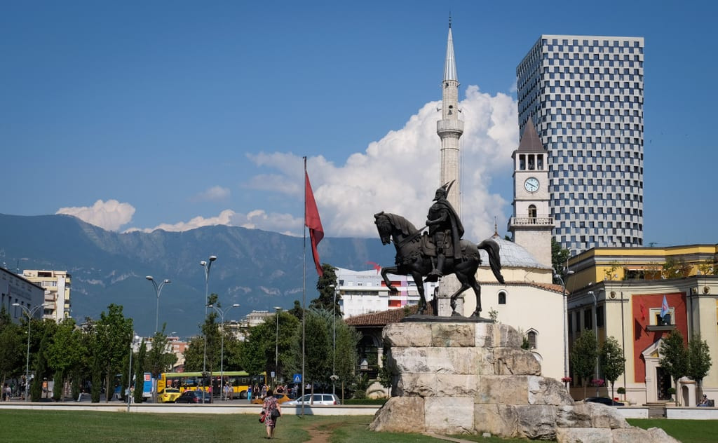 Tirana, with a statue of a man on a horse, a mosque with a minaret, a giant blood-red flag, a tall skyscraper, and mountains in the background.