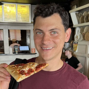 Nomadic Matt holding up a piece of pizza at a pizza shop in NYC