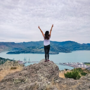 Một cô gái đứng trên mỏm đá nhìn ra hồ và núi non ở New Zealand. Xin visa làm việc kết hợp kỳ nghỉ ở New Zealand là một trong những công việc tốt nhất cho phép bạn đi du lịch mà không cần kinh nghiệm
