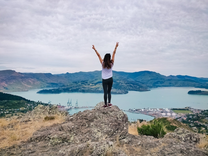 Một cô gái đứng trên mỏm đá nhìn ra hồ và núi non ở New Zealand. Xin visa làm việc kết hợp kỳ nghỉ ở New Zealand là một trong những công việc tốt nhất cho phép bạn đi du lịch mà không cần kinh nghiệm