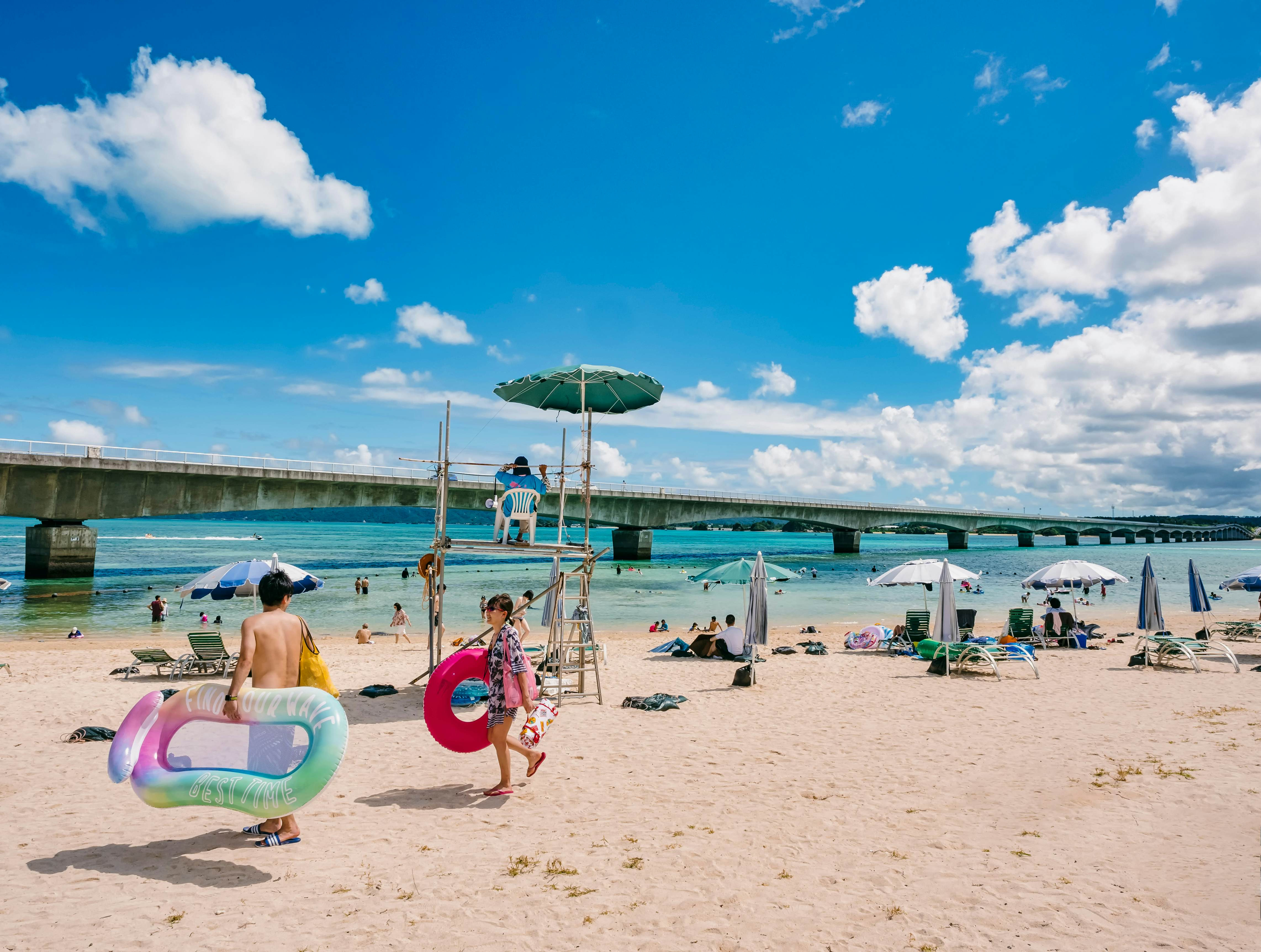 Du khách đi bộ qua Bãi biển Kouri trong kỳ nghỉ hè ở Okinawa, Nhật Bản, với bầu trời xanh trong vắt phía trên.